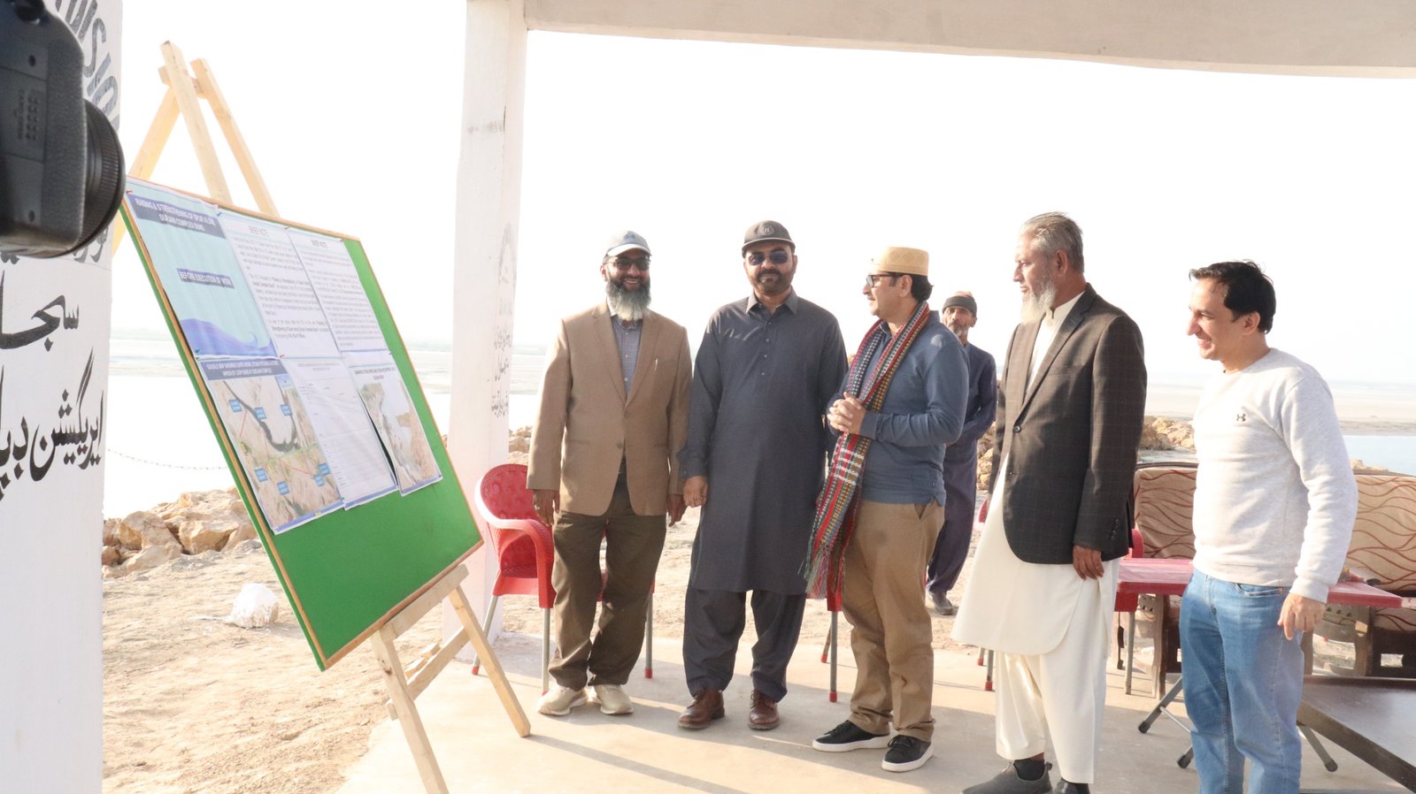 Bilal Anwar and colleagues under a shade structure for a coastal site briefing, a board of maps and photographs beside them