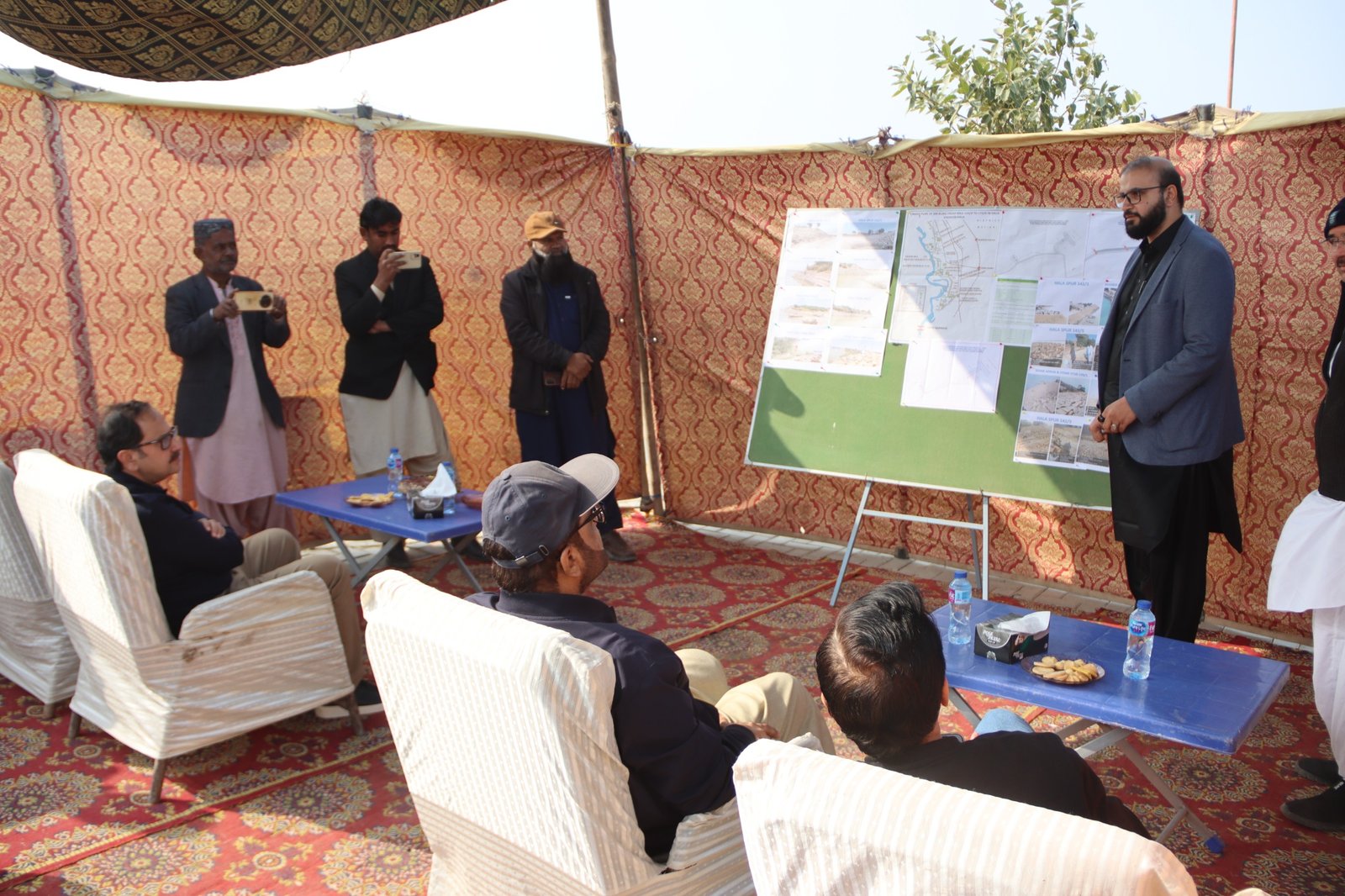 A colleague briefing a seated group from a board of maps and photographs inside a decorated tent
