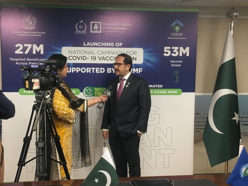 Bilal Anwar giving a television interview to camera in front of the backdrop for the national COVID-19 vaccination campaign supported by NDRMF